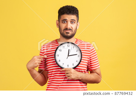 Look at time. Unhappy impatient bearded guy in striped t-shirt pointing big clock and looking displeased irritated, showing wall watches to hurry up. Indoor studio shot isolated on yellow background Look at time. Unhappy impatient bearded guy in striped t-shirt pointing big clock and looking displeased irritated, showing wall watches to hurry up. Indoor studio shot isolated on yellow background 87769432