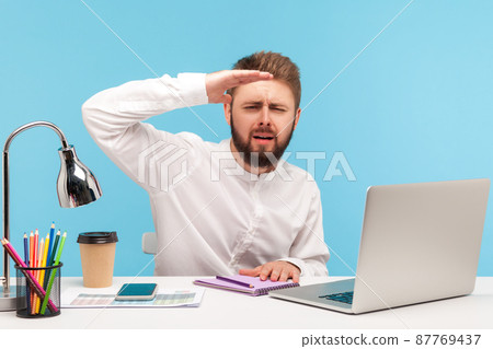 Ambitious bearded man office worker sitting workplace with laptop and looking far away with hand above eyes, searching with promotion, exploring future. Indoor studio shot isolated on blue background Ambitious bearded man office worker sitting workplace with laptop and looking far away with hand above eyes, searching with promotion, exploring future. Indoor studio shot isolated on blue background 87769437