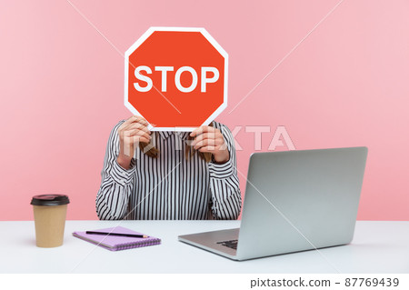 Female office worker in striped shirt sitting at workplace hiding face behind red stop traffic sign avoiding conflicts, afraid of workplace bullying. Indoor studio shot isolated on pink background 87769439