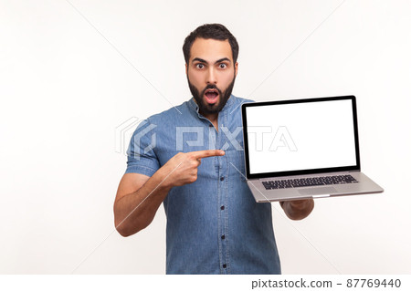 Surprised man pointing finger at empty display of laptop looking at camera with big eyes and opened mouth, shocked with advertisement. Indoor studio shot isolated on white background 87769440