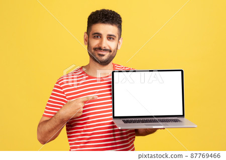 Smiling positive man with beard in striped t-shirt pointing finger at laptop with empty white display, recommending online education courses. Indoor studio shot isolated on yellow background Smiling positive man with beard in striped t-shirt pointing finger at laptop with empty white display, recommending online education courses. Indoor studio shot isolated on yellow background 87769466
