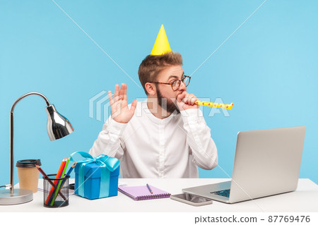 Funny man in party cap blowing into party whistle looking at laptop display, congratulating with holiday online via web camera, gift box lying on desk. Indoor studio shot isolated on blue background 87769476