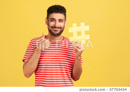 Cheerful satisfied man with beard in striped t-shirt showing thumbs up like gesture holding yellow hashtag sign, satisfied with target advertisement. Indoor studio shot isolated on yellow background 87769484