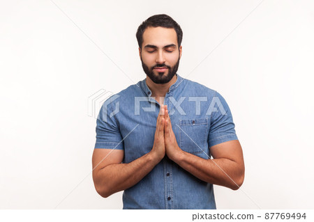 Calm and relaxed bearded man in blue shirt pressing hands together and smiling, meditating and opening his mind, standing with closed eyes. Indoor studio shot isolated on white background 87769494