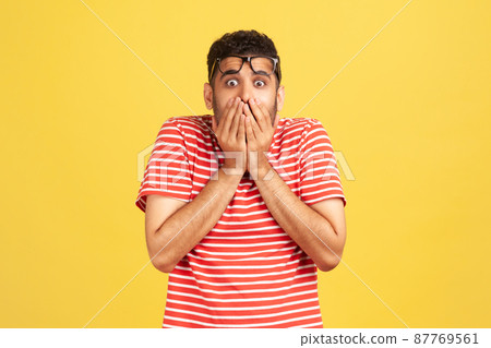 Shocked scared man in striped t-shirt closing mouth with hands, confused and depressed with information, looking with astonishment and fear, privacy. Indoor studio shot isolated on yellow background Shocked scared man in striped t-shirt closing mouth with hands, confused and depressed with information, looking with astonishment and fear, privacy. Indoor studio shot isolated on yellow background 87769561