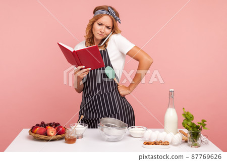 Puzzled young adult woman baker taking phone and asking help with preparing pastry, holding notebook in hands, surrounded with bakery ingredients. Indoor studio shot isolated on pink background. Puzzled young adult woman baker taking phone and asking help with preparing pastry, holding notebook in hands, surrounded with bakery ingredients. Indoor studio shot isolated on pink background. 87769626