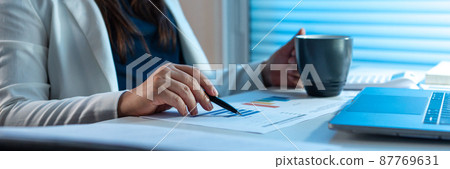 Overwork Concept The office woman who works at her desk pointing her pen on the paper with right hand and holding a cup with her left hand 87769631