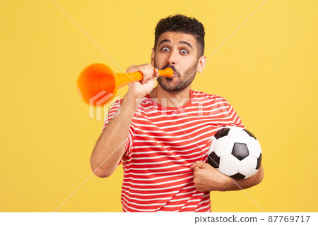 Very happy cheerful man with beard in striped t-shirt blowing in big bullhorn holding football ball, celebrating championship beginning. Indoor studio shot isolated on yellow background Very happy cheerful man with beard in striped t-shirt blowing in big bullhorn holding football ball, celebrating championship beginning. Indoor studio shot isolated on yellow background 87769717
