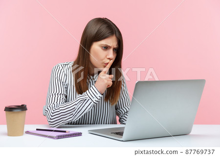 Please, keep secret. Concerned woman employee sitting at workplace shushing with silence gesture, talking on video call, online communication via laptop. Indoor studio shot isolated on pink background Please, keep secret. Concerned woman employee sitting at workplace shushing with silence gesture, talking on video call, online communication via laptop. Indoor studio shot isolated on pink background 87769737