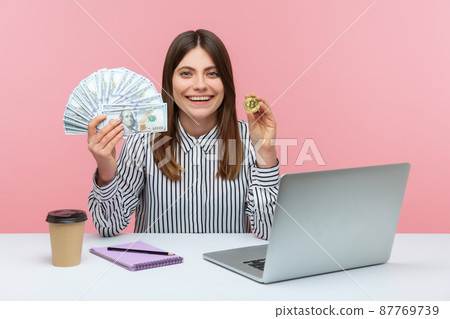 Positive woman office worker holding and showing fan of hundred dollar banknotes and bitcoin crypto coin looking at camera with toothy smile. Indoor studio shot isolated on pink background 87769739