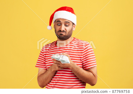 Unhappy confused bearded man in santa claus hat holding dollars in hands, greedy to buy holiday gifts. Indoor studio shot isolated on yellow background 87769758