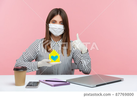 Work at home, stay safe. Woman employee sitting at workplace, wearing hygienic mask and protective gloves, holding paper house and showing thumbs up, remote job during coronavirus quarantine. indoor Work at home, stay safe. Woman employee sitting at workplace, wearing hygienic mask and protective gloves, holding paper house and showing thumbs up, remote job during coronavirus quarantine. indoor 87769800