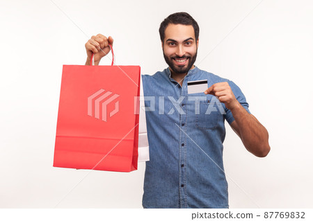 Happy satisfied man with beard holding and showing limitless credit card and shopping bags, pleased with great shopping, final sale. Indoor studio shot isolated on white background 87769832