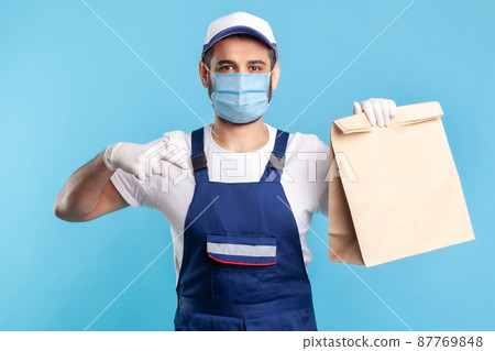Look at bag. Portrait of happy smiling handyman in overalls, mask and gloves pointing at ordered parcel. Courier delivering food in paper package, post mail services. isolated on blue background Look at bag. Portrait of happy smiling handyman in overalls, mask and gloves pointing at ordered parcel. Courier delivering food in paper package, post mail services. isolated on blue background 87769848
