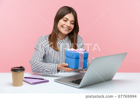 Cheerful happy woman siting at workplace showing gift box to laptop screen, talking on video call, celebrating birthday alone on self quarantine. Indoor studio shot isolated on pink background 87769994