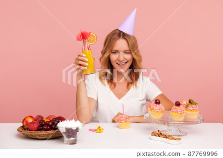 Happy blonde young adult woman raising hand with cocktail and winking eye, looks at camera smiling, celebrating birthday alone, wearing party cone. Indoor studio shot isolated on pink background. 87769996
