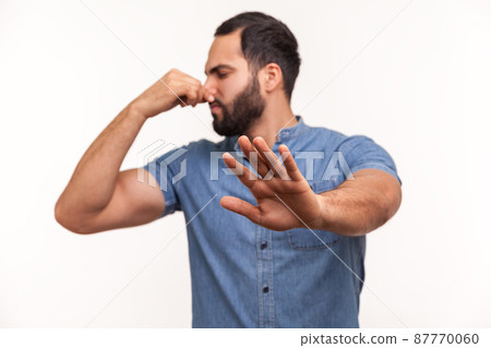Confused unhappy bearded man grabbing nose with fingers and stretching hand out feeling disgusted unpleasant smell. Indoor studio shot isolated on white background Confused unhappy bearded man grabbing nose with fingers and stretching hand out feeling disgusted unpleasant smell. Indoor studio shot isolated on white background 87770060