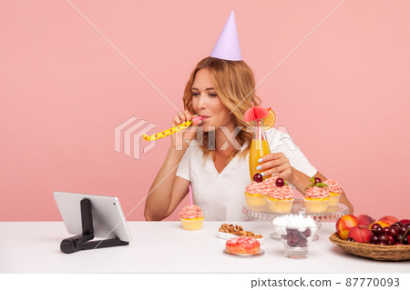 Festive blonde young adult woman wearing party cone and blowing paper horn, blogger celebrating birthday with cocktail and followers online. Indoor studio shot isolated on pink background. Festive blonde young adult woman wearing party cone and blowing paper horn, blogger celebrating birthday with cocktail and followers online. Indoor studio shot isolated on pink background. 87770093