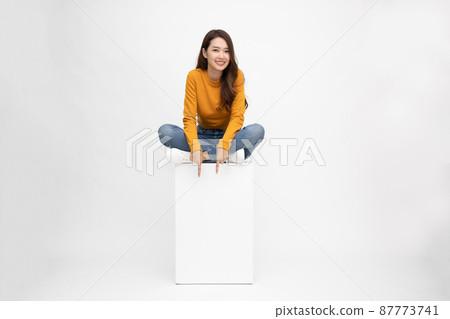 Smiling young Asian woman sitting on white box and pointing down empty copy space isolated on white background 87773741