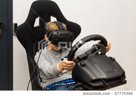 The boy plays on the racing simulator in glasses of a virtual reality The boy plays on the racing simulator in glasses of a virtual reality 87774366
