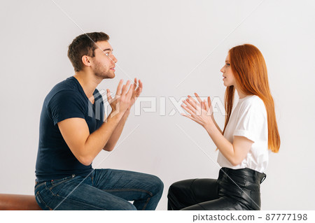 Studio shot of angry young couple quarreling, actively gesturing with hands, talking and shouting at each other on white isolated background. Concept of relationship crisis. 87777198