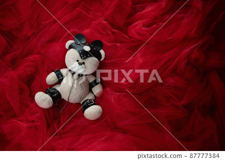 a toy teddy bear dressed in leather belts and a mask, an accessory for BDSM games against a beautiful red fabric 87777384