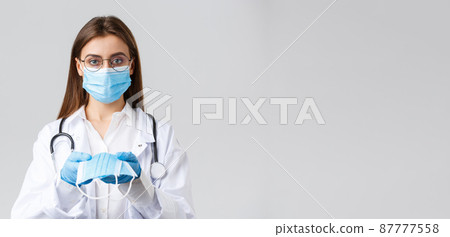 Covid-19, preventing virus, clinic, healthcare workers and quarantine concept. Young doctor in medical mask and gloves, white scrubs, giving face mask protection to patients, look serious 87777558