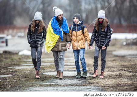 Woman holds Ukrainian flag as she flees the conflict with Russia with three kids in a derelict surroundings. Woman holds Ukrainian flag as she flees the conflict with Russia with three kids in a derelict surroundings. 87781726