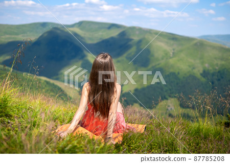 Young happy woman traveler in red dress resting on green grassy hillside on a windy day in summer mountains enjoying view of nature. Young happy woman traveler in red dress resting on green grassy hillside on a windy day in summer mountains enjoying view of nature. 87785208