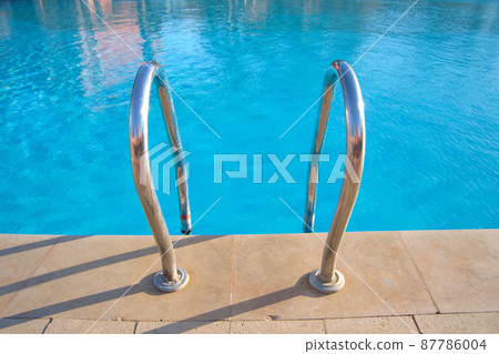 Close up of swimming pool stainless steel handrail descending into tortoise clear pool water. Accessibility of recreational activities concept. Close up of swimming pool stainless steel handrail descending into tortoise clear pool water. Accessibility of recreational activities concept. 87786004
