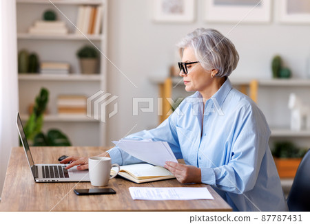 Mature businesswoman working at laptop, reading documents and take notes in home office 87787431
