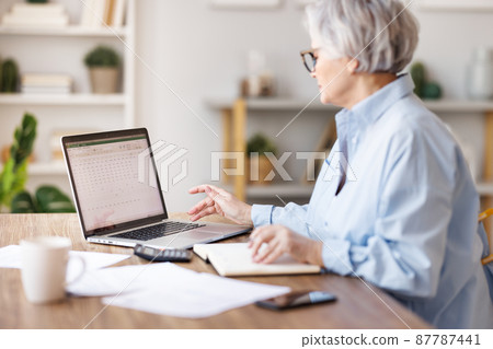 Mature businesswoman working at laptop, reading documents and take notes in home office 87787441