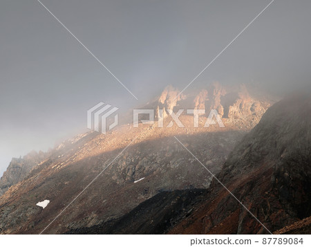 Atmospheric ghostly landscape with fuzzy silhouettes of sharp rocks in low clouds. Dramatic view to large mountains blurred in rain haze in gray low clouds. Diagonal slope of the mountain 87789084