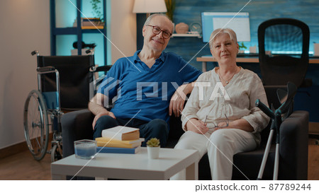 Portrait of retired couple with disability sitting on couch Portrait of retired couple with disability sitting on couch 87789244