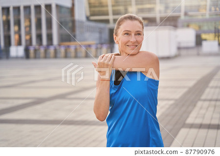 Portrait of attractive middle aged woman in sportswear stretching her arms, standing outdoors, getting ready for workout in the city 87790976