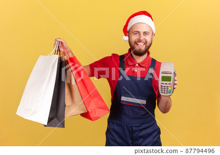 Positive smiling male courier in uniform and santa claus hat holding payment terminal and paper shopping bags, easy express order and delivery. Indoor studio shot isolated on yellow background Positive smiling male courier in uniform and santa claus hat holding payment terminal and paper shopping bags, easy express order and delivery. Indoor studio shot isolated on yellow background 87794496