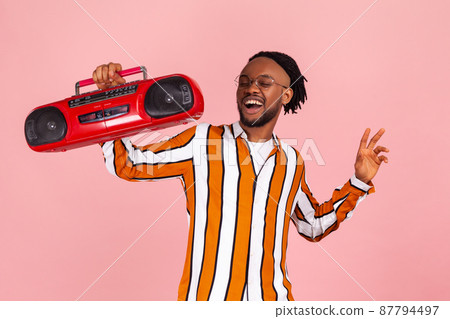 Happy bearded afro-american man music lover with dreadlocks in eyeglasses and stylish striped shirt enjoying music, holding red tape recorder in hand. Indoor studio shot isolated on pink background Happy bearded afro-american man music lover with dreadlocks in eyeglasses and stylish striped shirt enjoying music, holding red tape recorder in hand. Indoor studio shot isolated on pink background 87794497