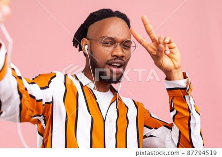 Successful excited african man blogger with beard and dreadlocks in trendy striped shirt showing v gesture posing at camera, making selfie. Indoor studio shot isolated on pink background 87794519