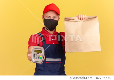 Courier in uniform and black protective mask showing paper bag and holding payment terminal, fast delivery and contactless payments on quarantine. Indoor studio shot isolated on yellow background 87794550
