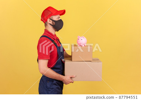 Profile portrait courier in uniform and protective mask holding cardboard parcels with piggy bank on top, save money ordering delivery, advertising. Indoor studio shot isolated on yellow background 87794551