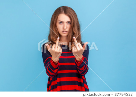 Portrait of crazy angry woman wearing striped casual style sweater showing middle finger, rude gesture, face full of hatred and resentment. Indoor studio shot isolated on blue background. 87794552