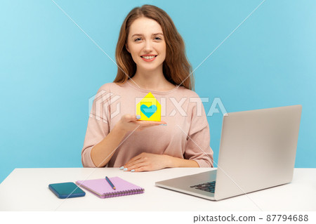 Insurance agency. Cheerful pretty woman holding paper house with heart inside and smiling, real estate agent sitting at desk with laptop, home office. indoor studio shot isolated on blue background 87794688