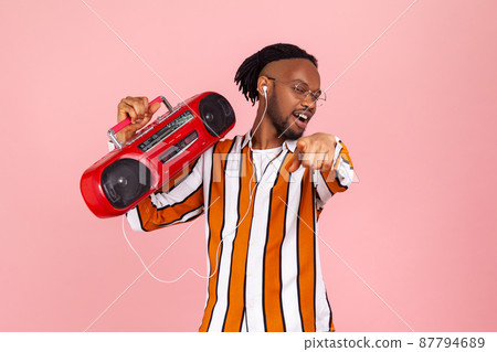 Positive satisfied african man with dreadlocks in striped shirt pointing finger on you holding red record player listening to music in earphones Indoor studio shot isolated on pink background 87794689