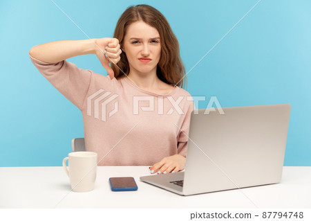 I don't like. Displeased woman employee showing thumbs down, dislike gesture, expressing disapproval, sitting at workplace with laptop, home office. indoor studio shot isolated on blue background I don't like. Displeased woman employee showing thumbs down, dislike gesture, expressing disapproval, sitting at workplace with laptop, home office. indoor studio shot isolated on blue background 87794748