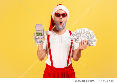 Shocked amazed man in sunglasses and santa claus red costume holding and showing payment terminal and dollar banknotes, choosing cashless payments. Indoor studio shot isolated on yellow background Shocked amazed man in sunglasses and santa claus red costume holding and showing payment terminal and dollar banknotes, choosing cashless payments. Indoor studio shot isolated on yellow background 87794750
