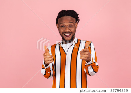 Surprised bemused african man with beard and dreadlocks in striped shirt pointing fingers at you and looking with astonishment, unbelievable. Indoor studio shot isolated on pink background Surprised bemused african man with beard and dreadlocks in striped shirt pointing fingers at you and looking with astonishment, unbelievable. Indoor studio shot isolated on pink background 87794859
