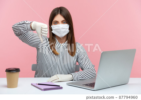 Displeased woman employee wearing hygienic mask and protective gloves during quarantine, showing thumbs down, dislike gesture. Working at home office in self-isolation, coronavirus outbreak. Indoor 87794960