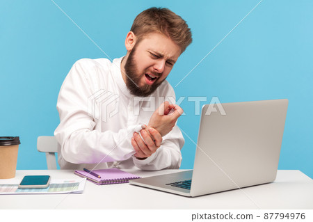 Bearded man office worker in white shirt grimacing feeling tingling, numbness and pain in wrist working on laptop, suffering carpal tunnel syndrome. Indoor studio shot isolated on blue background Bearded man office worker in white shirt grimacing feeling tingling, numbness and pain in wrist working on laptop, suffering carpal tunnel syndrome. Indoor studio shot isolated on blue background 87794976