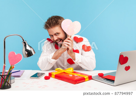 Playful bearded man sitting at workplace all covered with heart sticks and smiling, holding pink greeting card in heart shape, preparing gift. Indoor studio shot isolated on blue background 87794997