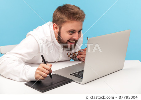 Curious man creative designer using graphic tablet and typing keyboard laptop, working on professional digital art equipment to draw sketch. Indoor studio shot isolated on blue background. Curious man creative designer using graphic tablet and typing keyboard laptop, working on professional digital art equipment to draw sketch. Indoor studio shot isolated on blue background. 87795004
